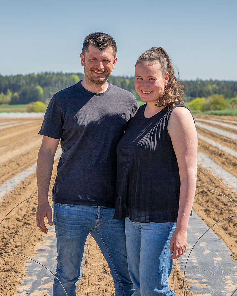 Ein Pärchen mit schwarzen Oberteuilen und blauen Jeans stehen lächelnd nah beieinander an einem sonnigen Frühlingstag auf einem Feld, an dem Kartoffeln gepflanzt wurden. Im Hintergrund sind weitere grüne Felder zu erkennen und Wald. Der Himmel ist strahlend Blau.