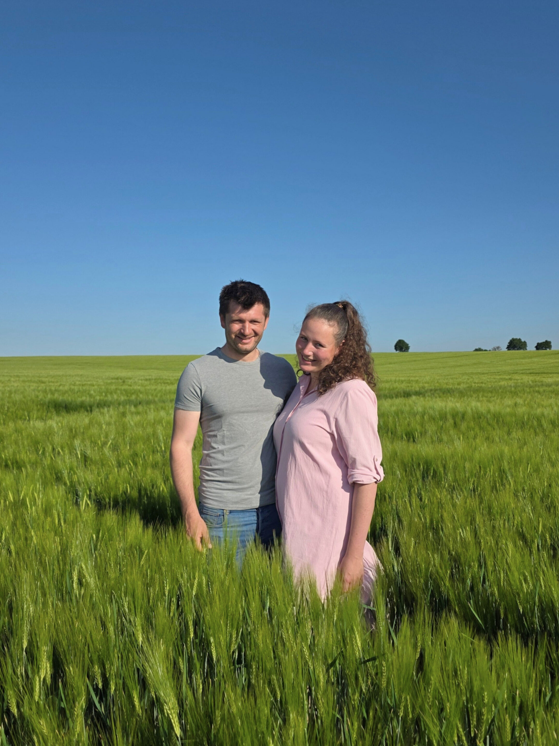 25.05.2026 _ Köglmaiers (3) Ein Pärchen mit schwarzen Oberteuilen und blauen Jeans stehen lächelnd nah beieinander an einem sonnigen Frühlingstag auf einem Feld, an dem Kartoffeln gepflanzt wurden. Im Hintergrund sind weitere grüne Felder zu erkennen und Wald. Der Himmel ist strahlend Blau.